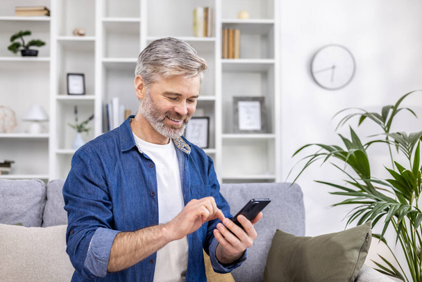 Mature adult man close-up at home smiling and using phone in living room sitting on sofa, person typing message and browsing web page. - Foto, Imagem