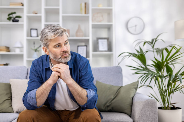 Adult mature man pensive sitting alone at home on sofa close up, gray haired person thinks looks towards window, depressed sad. - Fotoğraf, Görsel