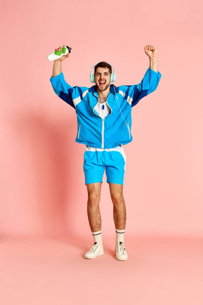 Positive active young man in uniform, headphones and with water bottle posing against pink studio background. Energy. Concept of sportive lifestyle, emotions, facial expression. Ad - Photo, image