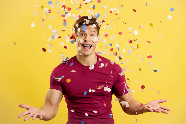 Hombre alegre rodeado de confeti volando en el aire en estudio con fondo amarillo - Foto, Imagen