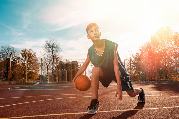 Sports and basketball. A young teenager in a green tracksuit playing basketball, leading the ball. Blue sky in the background and a sports ground in the background. Sunlight. Copy space. - Фото, изображение