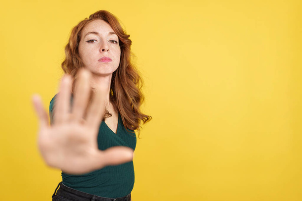 Рыжая женщина жестом запрет с рукой в студии с желтым фоном - Фото, изображение