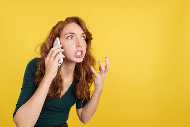 Mujer pelirroja enojada hablando con el móvil en estudio con fondo amarillo - Foto, imagen