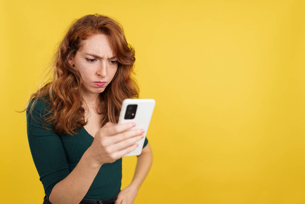 Bezorgde roodharige vrouw met behulp van een mobiele telefoon in de studio met gele achtergrond - Foto, afbeelding