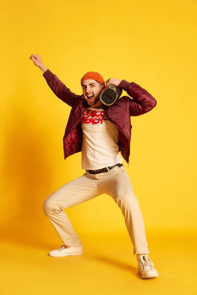 Portrait of handsome positive young man posing in stylish casual clothes with music player against yellow studio background. Concept of emotions, male fashion, lifestyle, facial expression. Ad - Фото, изображение