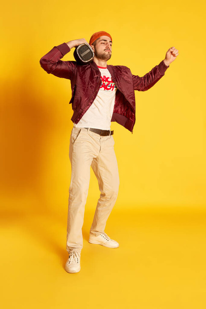 Portrait of cheerful stylish young man in casual clothes posing with music player on shoulders against yellow studio background. Concept of emotions, male fashion, lifestyle, facial expression. Ad - Photo, image