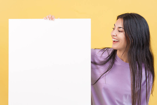 Happy caucasian woman looking and holding a blank panel in studio with yellow background - Photo, Image