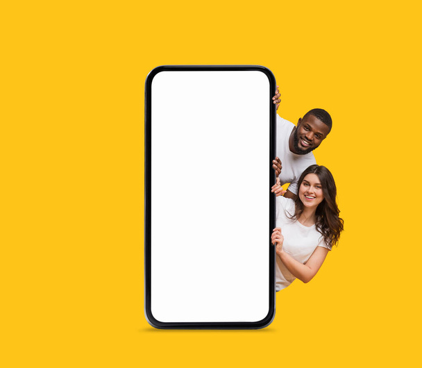 Mockup móvil. Pareja Interracial feliz asomándose detrás de un gran teléfono inteligente en blanco mientras está de pie aislado sobre el fondo amarillo, el hombre alegre y la mujer copian el espacio para el diseño de aplicaciones o sitios web, Collage - Foto, imagen