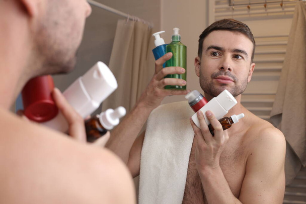 portret przystojnego młodzieńca z butelką środków higienicznych przed lustrem w łazience - Zdjęcie, obraz