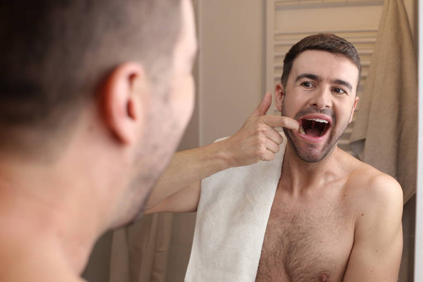 portret van knappe jongeman controleren zijn tanden in de voorkant van spiegel in de badkamer - Foto, afbeelding
