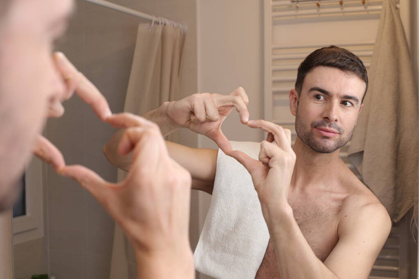 retrato de joven guapo mostrando la forma del corazón con las manos delante del espejo en el baño - Foto, Imagen
