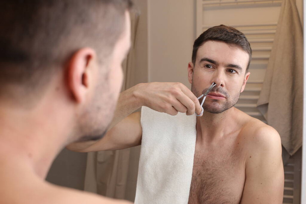 portrait of handsome young man trimming his nose hair in front of mirror in bathroom - Photo, Image