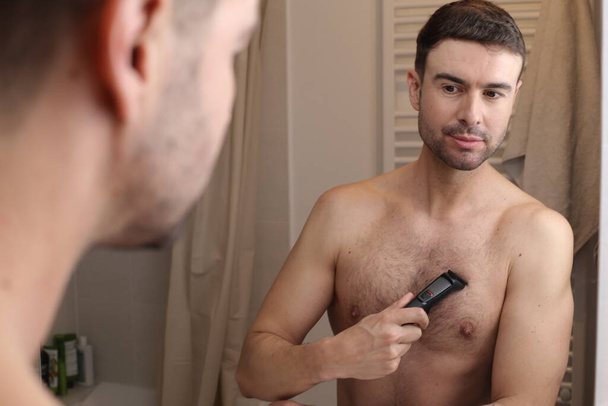 portrait of handsome young man trimming his chest hair with electric trimmer front of mirror in bathroom - Photo, Image