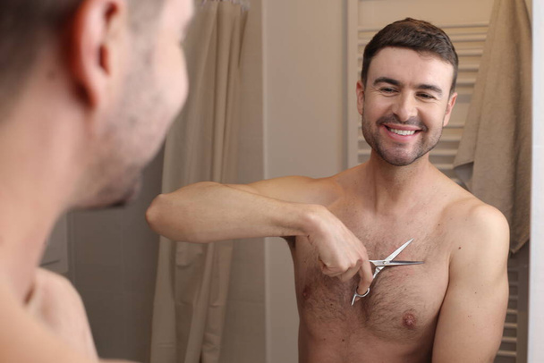 портрет красивого молодого чоловіка, який обрізає волосся на грудях ножицями перед дзеркалом у ванній
 - Фото, зображення