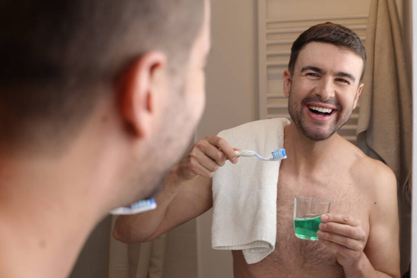 portrét pohledného mladého muže se zubním kartáčkem a ručníkem před zrcadlem v koupelně - Fotografie, Obrázek