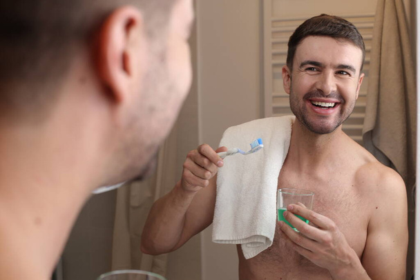 portret van knappe jongeman met tandenborstel en handdoek voor spiegel in badkamer - Foto, afbeelding