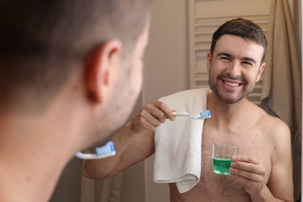 portrait of handsome young man with toothbrush and towel in front of mirror in bathroom - Foto, afbeelding