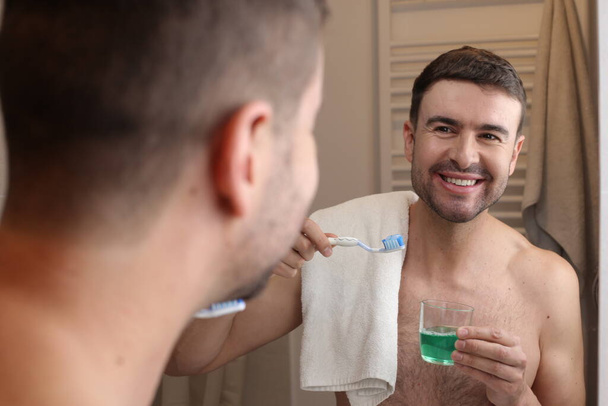 портрет красивого молодого человека с зубной щеткой и полотенцем перед зеркалом в ванной комнате - Фото, изображение