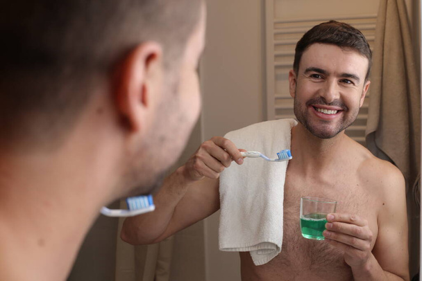 Porträt eines hübschen jungen Mannes mit Zahnbürste und Handtuch vor dem Spiegel im Badezimmer - Foto, Bild