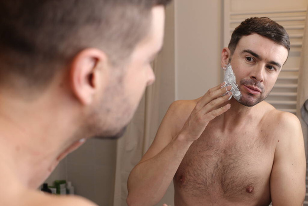 portrait of handsome young man applying shaving foam in front of mirror in bathroom - Фото, изображение