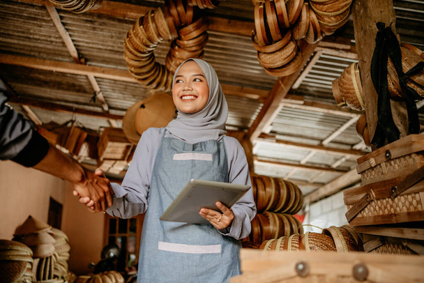 beautiful young muslim woman shake hand with customer in her woven bamboo product workshop - Фото, зображення