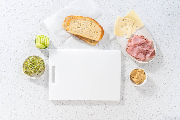 Επίπεδη. Συνάθροιση ζαμπόν, αγγούρι, και φυτρώνουν σάντουιτς στο λευκό πίνακα κοπής. - Φωτογραφία, εικόνα