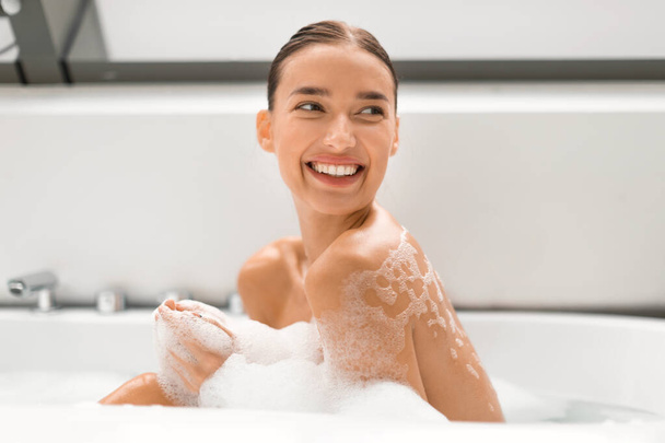 Happy Lady tomando banho relaxante lavando o corpo sentado na banheira cheia de bolhas de espuma no banheiro moderno em casa, sorrindo olhando para o lado. Rotina de beleza e conceito de relaxamento - Foto, Imagem