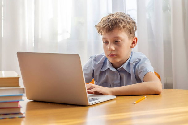 Thoughtful schoolboy studying online on laptop at desk with books and stationery - Photo, Image