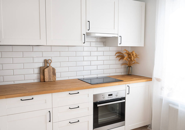 Accogliente cucina bianca con piano di lavoro in legno, fiori e taglieri in un interno minimalista con luce solare durante il giorno. Vista diagonale. - Foto, immagini