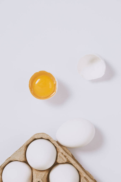 Cracked egg half with a yolk inside near the egg tray with eggs on bright background. Selective focus. Concept scene. Top view - Zdjęcie, obraz
