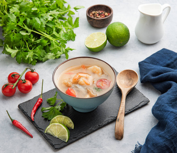 Tom yam soup with shrimp and chili pepper in a blue bowl on a light background with fresh vegetables and herbs. Traditional Asian food. Thai dish concept. - Fotografie, Obrázek