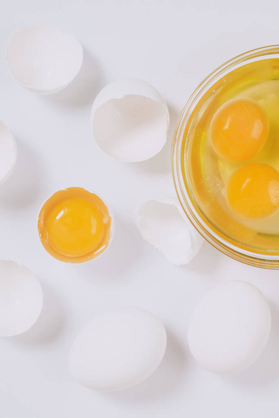 Whole white eggs and cracked white eggs half with a yolk inside near the glass bowl with egg yolks and egg whites on bright background. Close up, selective focus. Top view. - Foto, Imagem