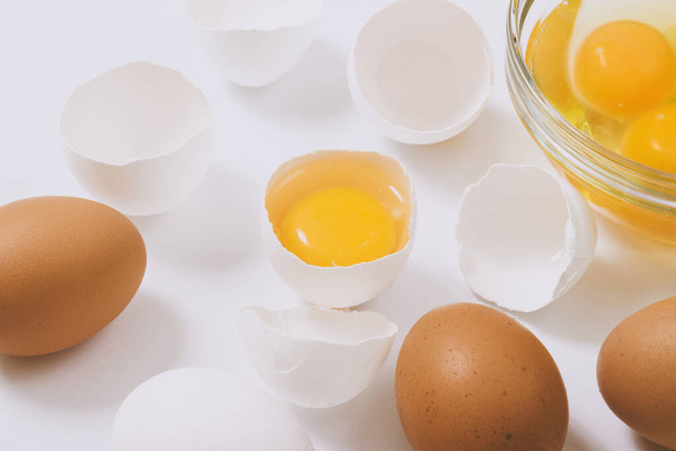 Celá hnědá vajíčka a popraskaná bílá vajíčka s žloutkem uvnitř u skleněné mísy s bílky a žloutky na jasném pozadí. Zavřít, selektivní zaměření. - Fotografie, Obrázek