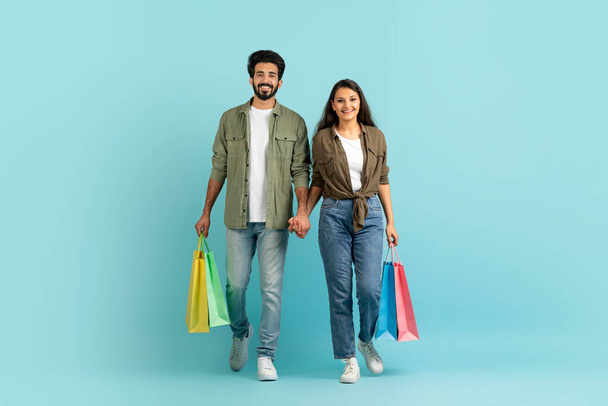 Happy emotional millennial multiracial couple holding colorful shopping bags, pretty indian lady walking with arab guy, isolated on blue studio background. Sales, retail, shopping concept - Fotoğraf, Görsel