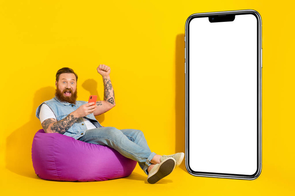 Photo of lucky cool man wear jeans waistcoat sitting beanbag rising fist modern device empty space isolated yellow color background. - Foto, Bild