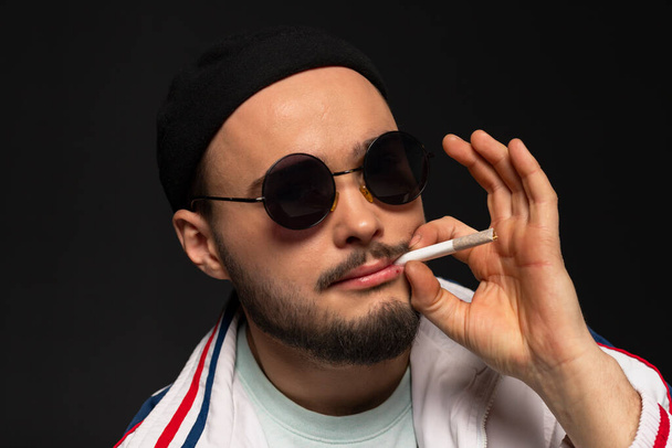 Close-up portrait of a oung, tough guy from the streets dressed in a tracksuit and round sunglasses smoking a cigarette. Urban street culture. isolated on a black background. - Photo, Image