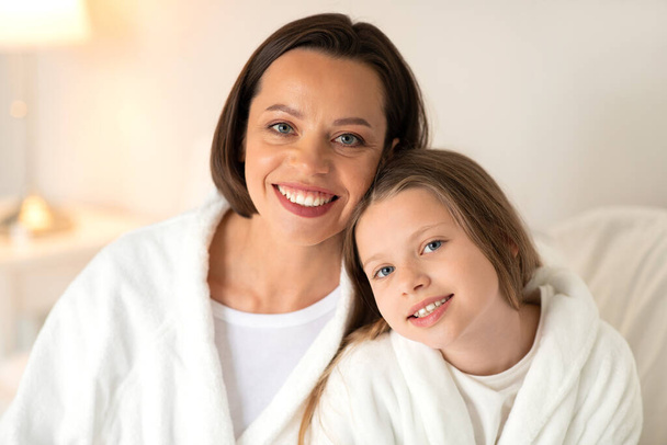 Smiling european millennial female, little girl in bathrobes hugs, sit on soft bed, enjoy rest together, free time, weekend in white bedroom interior. Good morning, comfort, relationship and love - Photo, Image