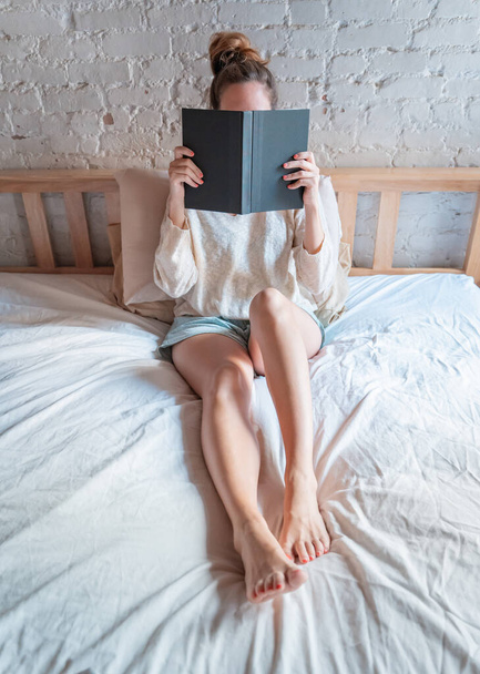 ベッドの中で怠惰な時間。認識できない若い女性はベッドの上に座って朝リラックスし、居心地の良いベッドルームで本を読んでください。スペースの垂直写真をコピーします。高品質の写真 - 写真・画像