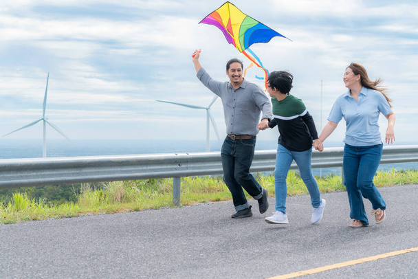 Progressive happy carefree family vacation concept. Young parents mother father and son run along and flying kite together on road with natural scenic on mountain and wind turbine background. - Foto, afbeelding