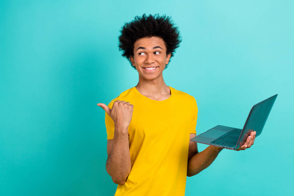 Photo of cheerful guy afro hairstyle dressed yellow t-shirt look directing empty space hold laptop isolated on turquoise color background. - Foto, imagen