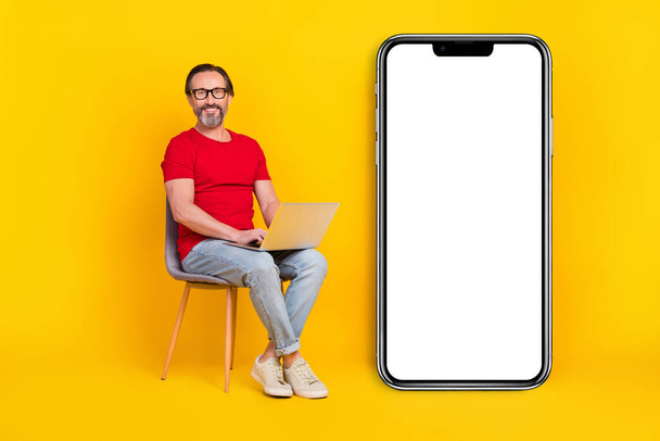 Photo of cute cheerful guy pensioner dressed red t-shirt glasses writing gadget empty space isolated yellow color background. - Foto, imagen