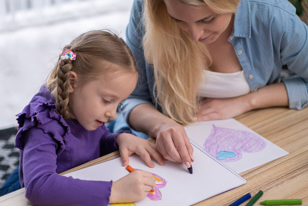 matka a dcera kreslí a barvísrdeční tvary na papíře. Máma pomáhá dívce naučit se malovat pastelkami - Fotografie, Obrázek