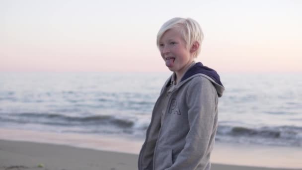 Ein Schuljunge mit weißem Haar streckt seine Zunge am Strand am Meer bei Sonnenuntergang aus. - Filmmaterial, Video