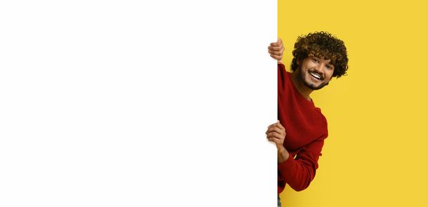 Fröhlich positiver gutaussehender junger indischer Typ, der von hinten horizontal leere weiße Werbetafel betrachtet und in die Kamera lächelt, gelber Studiohintergrund, Kopierraum, Panorama - Foto, Bild