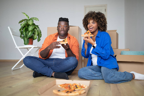 Giovani sposi neri che si riposano nel giorno del trasloco, mangiano pizza da asporto e guardano la TV mentre sono seduti sul pavimento tra scatole di cartone, coppia afroamericana che celebra la delocalizzazione in una nuova casa - Foto, immagini