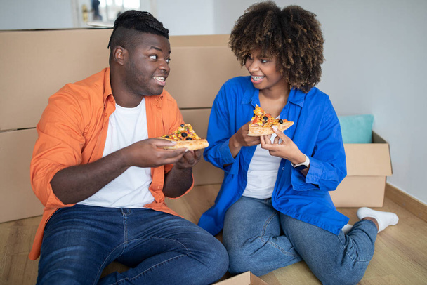 Junges schwarzes Paar isst Pizza und entspannt sich am Tag des Umzugs auf dem Boden, romantische afroamerikanische Ehepartner sitzen zwischen Pappkartons, essen zu Mittag und feiern Umzug, Nahaufnahme - Foto, Bild