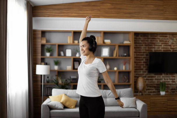 Attractive interracial woman in activewear listening music in wireless headphone during morning exercises at apartment. Happy young lady enjoying active and healthy time spending alone at home. - Фото, изображение