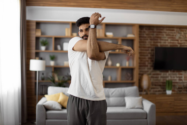 Handsome focused indian man stretching arms for bodybuilder fitness and muscle warm up at cozy living room. Wellness, sport activity and people concept. - Photo, image