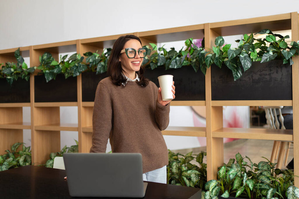 Happy European Female Entrepreneur Standing Near Desk With Laptop Computer Posing Smiling In Modern Office Indoor. Freelance Career And Successful Entrepreneurship Concept - Foto, Imagem