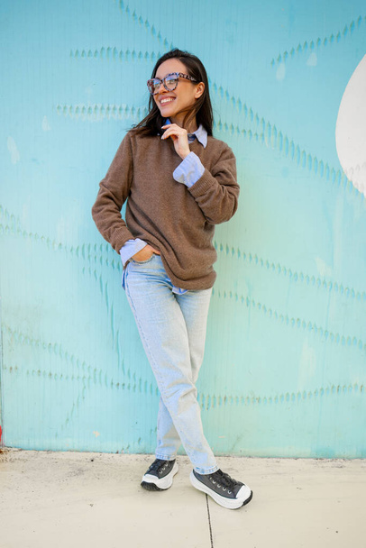 Happy woman in glasses outdoor on blue color background. Positive people concept. Smiling girl looking away, hands in pocket, dressed sweater and jeans. Vertical full length - Zdjęcie, obraz